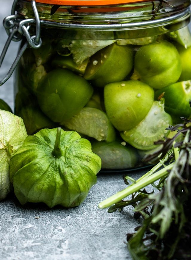 Einmachglas mit grünen Tomatillos in Sud, daneben eine frische Tomatillo mit papierartiger Hülle